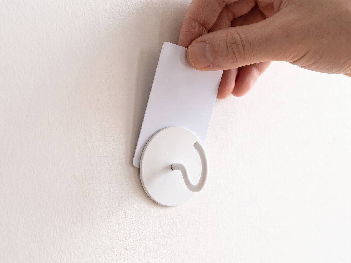 Eine Hand schiebt eine Plastikkarte von oben hinter die runde Platte eines weißen Klebehakens